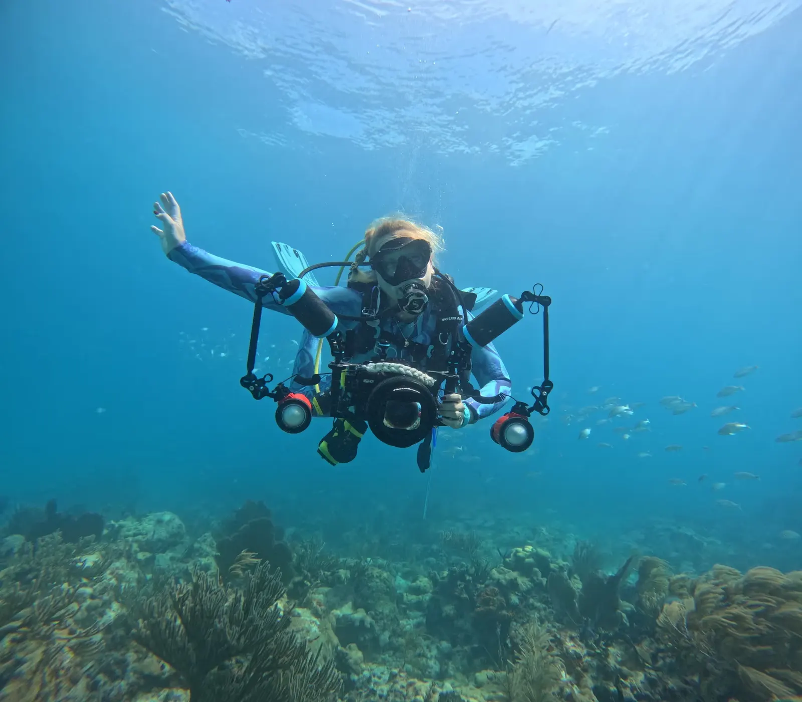 Scuba diver underwater holding an underwater camera rig with lights, surrounded by coral and fish.