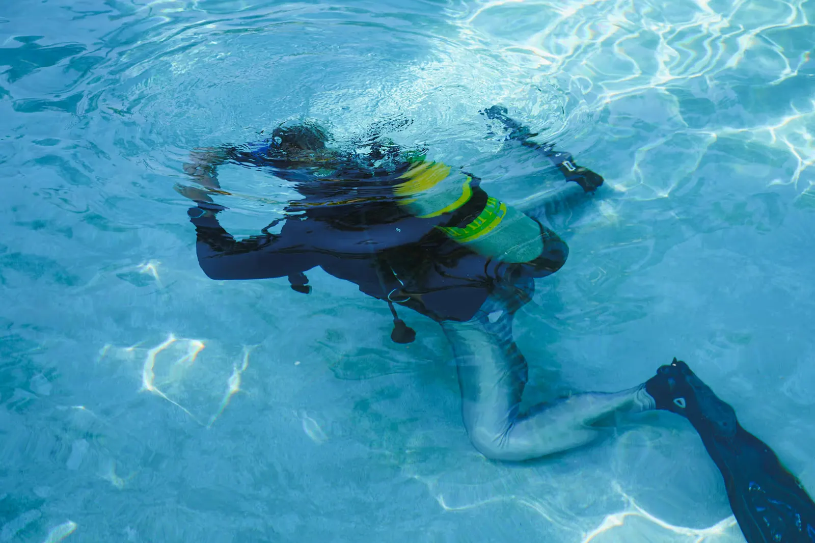 Scuba diver underwater in clear blue water wearing fins and carrying a yellow-green air tank.