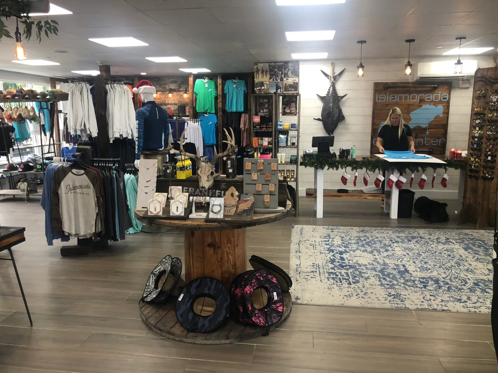 Interior of a retail store with clothing racks, a display table with jewelry and antlers, and a woman folding shirts behind a counter decorated with Christmas stockings and greenery.