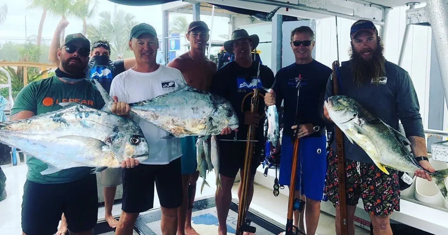 Group of seven people on a dive boat holding large permit fish and spearguns with Florida Keys Dive Center shirts visible after a successful spearfishing charter